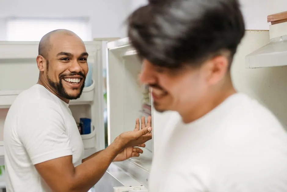 Two men in a kitchen laughing together near the refrigerator, enjoying a lighthearted moment.